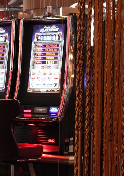 A pair of casino slot machines, each with detailed screens displaying various game outcomes and jackpot amounts. The chairs in front of the slot machines are dark and cushioned for comfort. The background features a hanging curtain made of golden ropes and other casino decorations are visible.